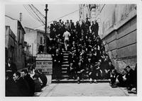 Large group of several dozen men seated on steps to a building, with sign reading BASE SECTION 64TH GENERAL HOSPITAL ENTRANCE