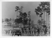Large group of soldiers lined up outside with gear; Jeep at left